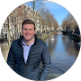 Man in a blue quilted jacket posing by a canal with trees and historic buildings in the background.
