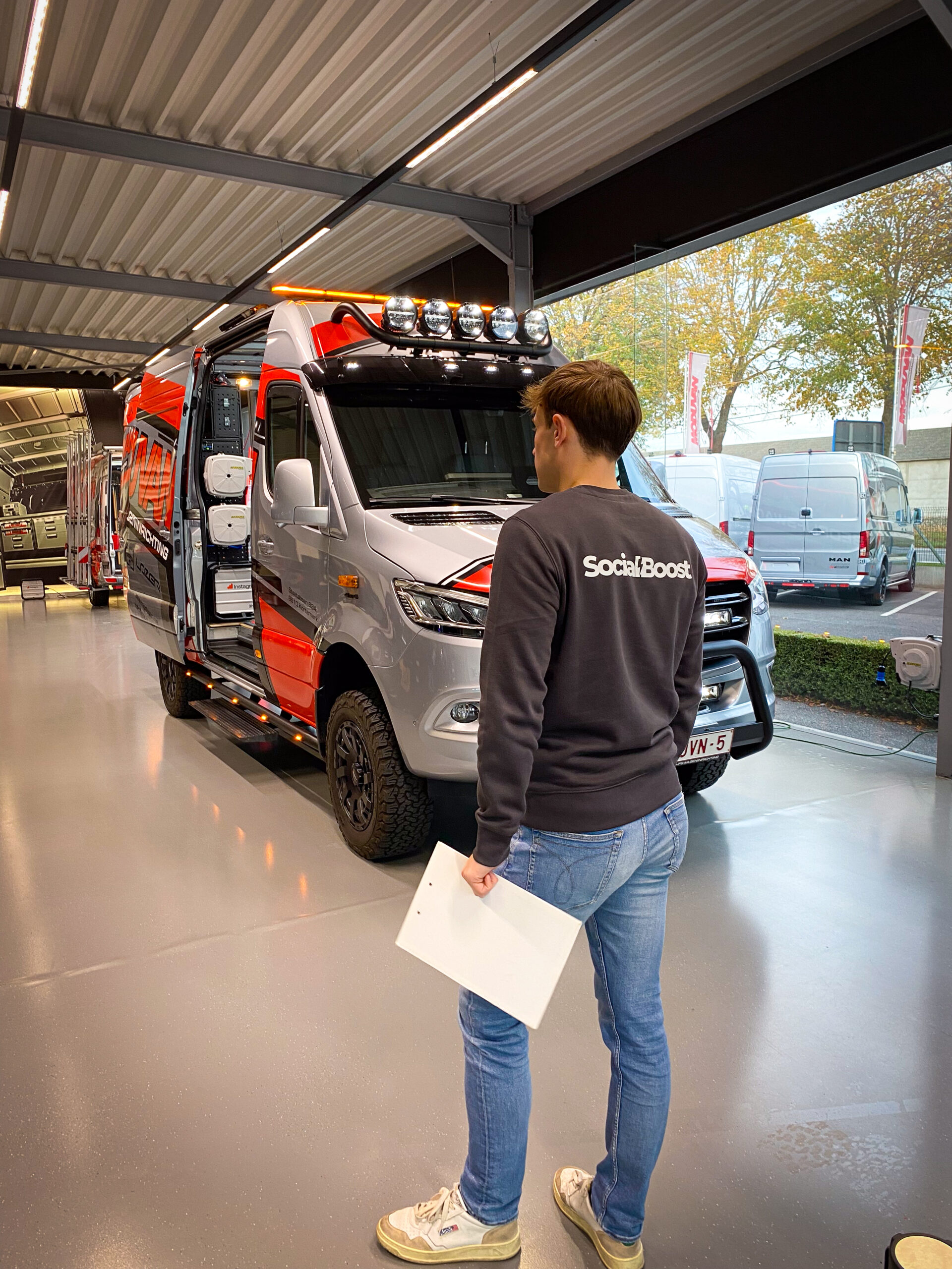 Man wearing a SocialBoost shirt stands next to a large red and silver van with roof-mounted lights inside a showroom.