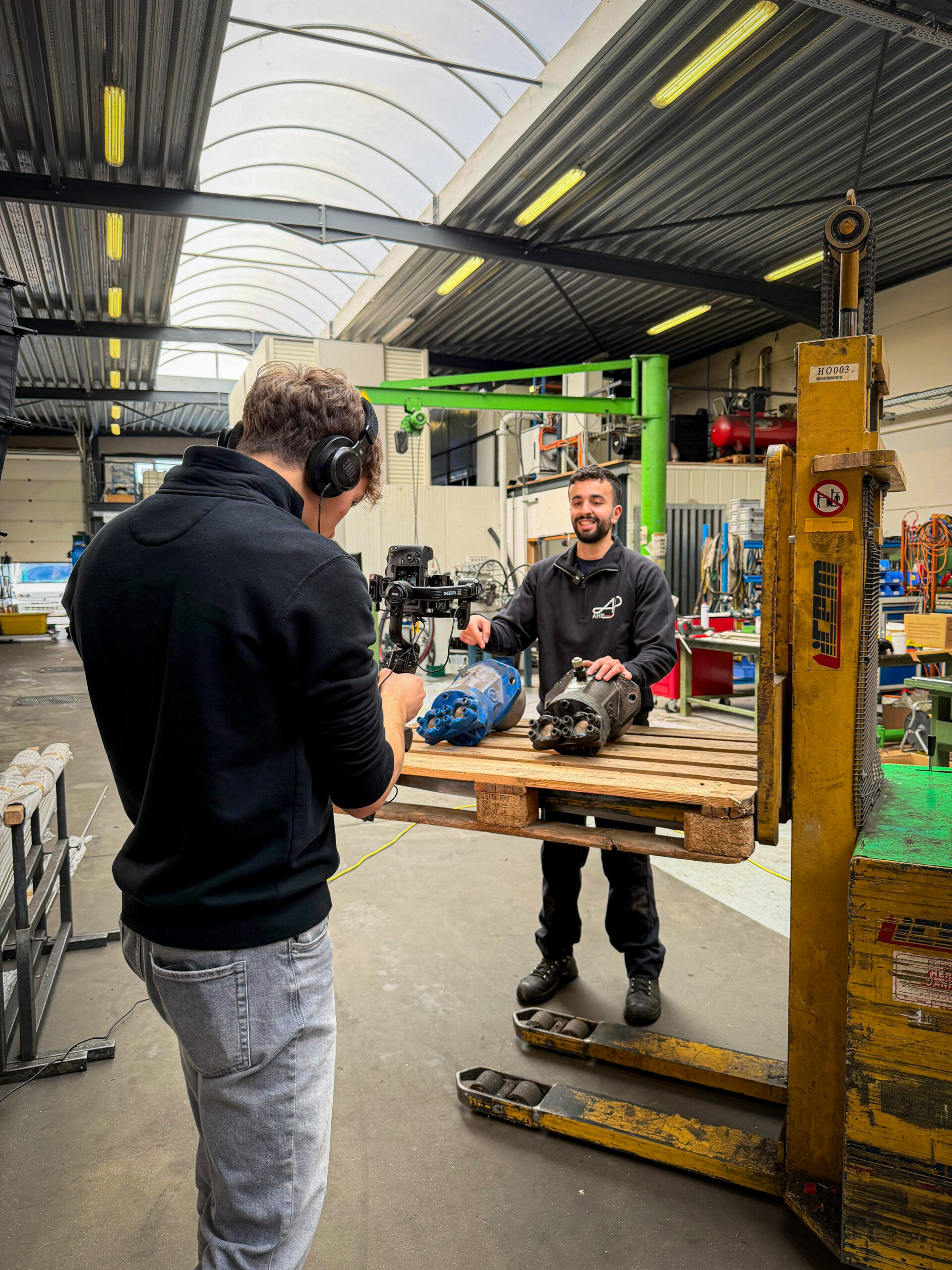 Two workers in an industrial workshop; one films a teammate explaining large mechanical parts on a wooden pallet, nearby a yellow pallet lift and tools lie around.
